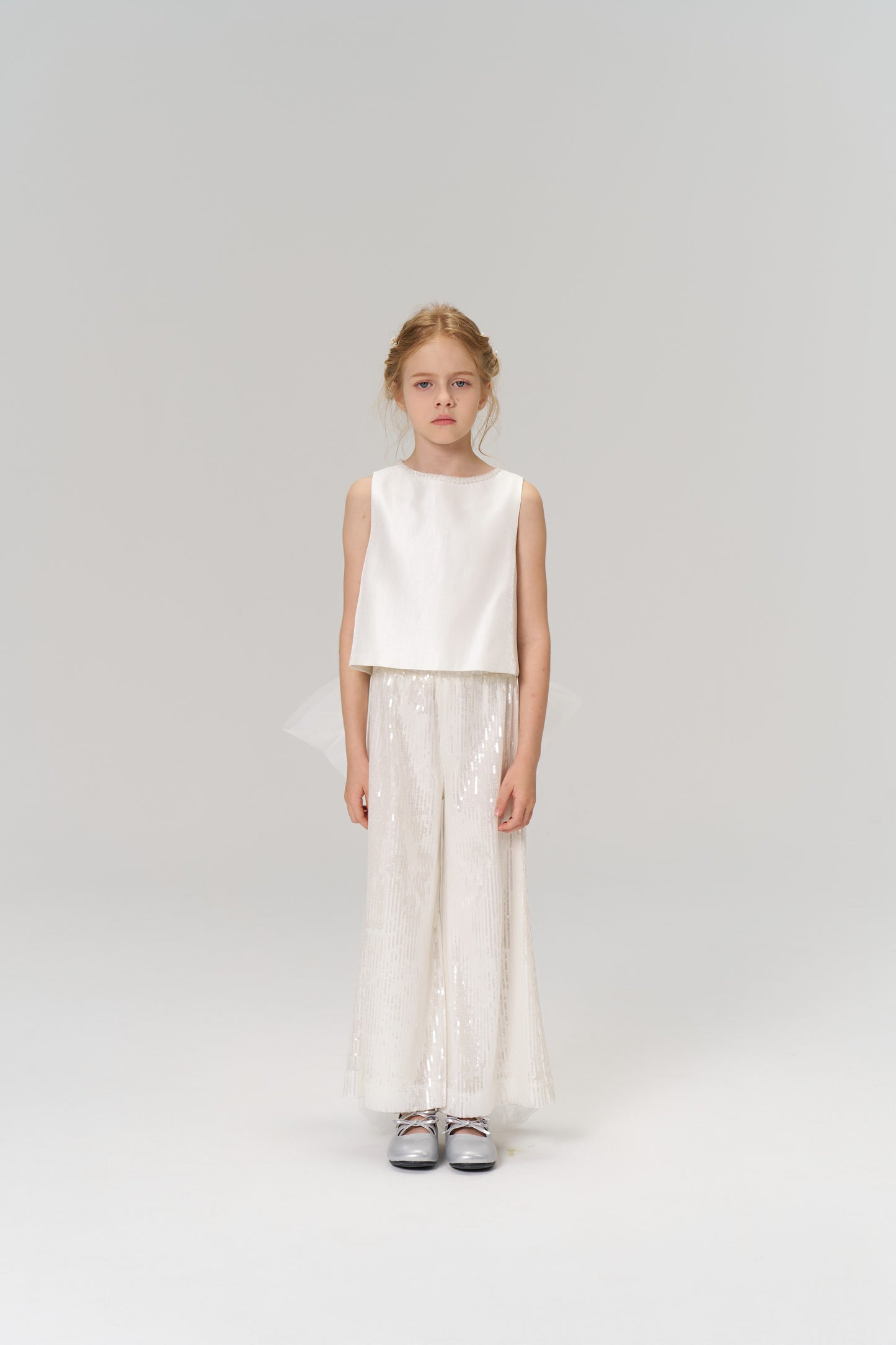 A young girl faces forward in the KIKI and JOJO Radiance Satin Crop Top with a hand-beaded pearl neckline, paired with white sequin wide-leg pants and silver shoes—an ideal look for holiday parties against a plain background.