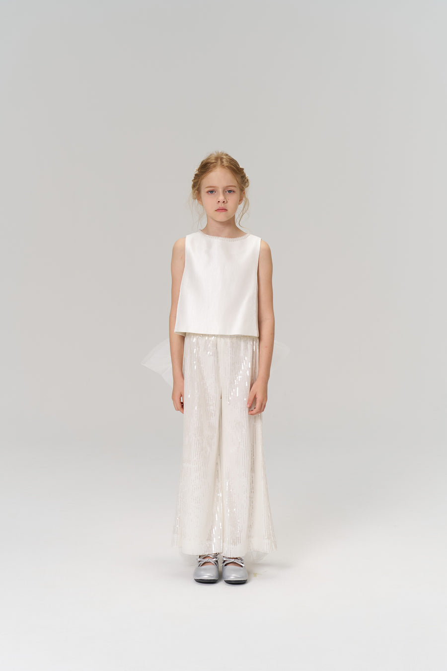 A young girl faces forward in the KIKI and JOJO Radiance Satin Crop Top with a hand-beaded pearl neckline, paired with white sequin wide-leg pants and silver shoes—an ideal look for holiday parties against a plain background.