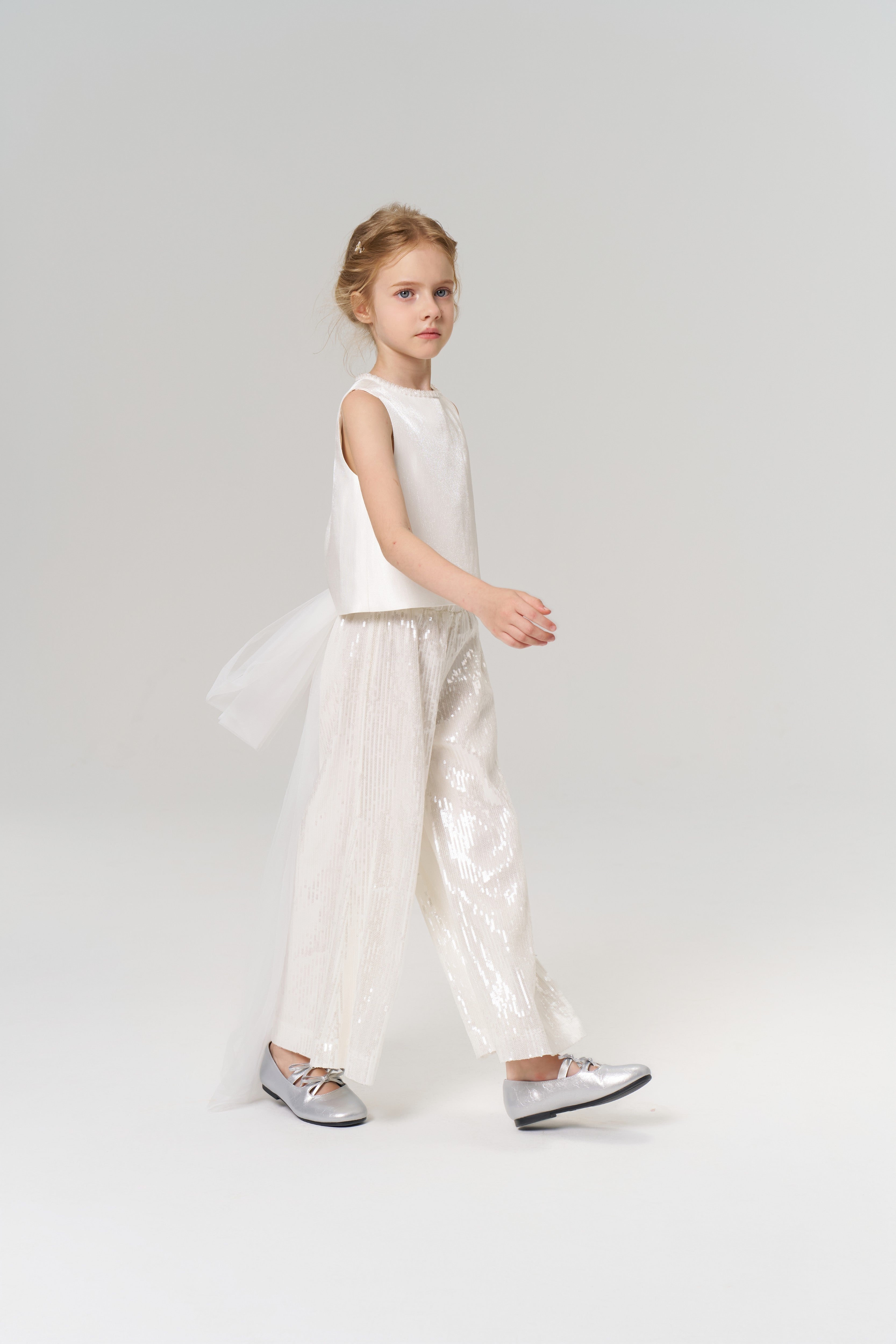 A young girl in a sleeveless white top and KIKI and JOJO Radiance Sequin Wide-Leg Pants with silver shoes walks across a plain light background.