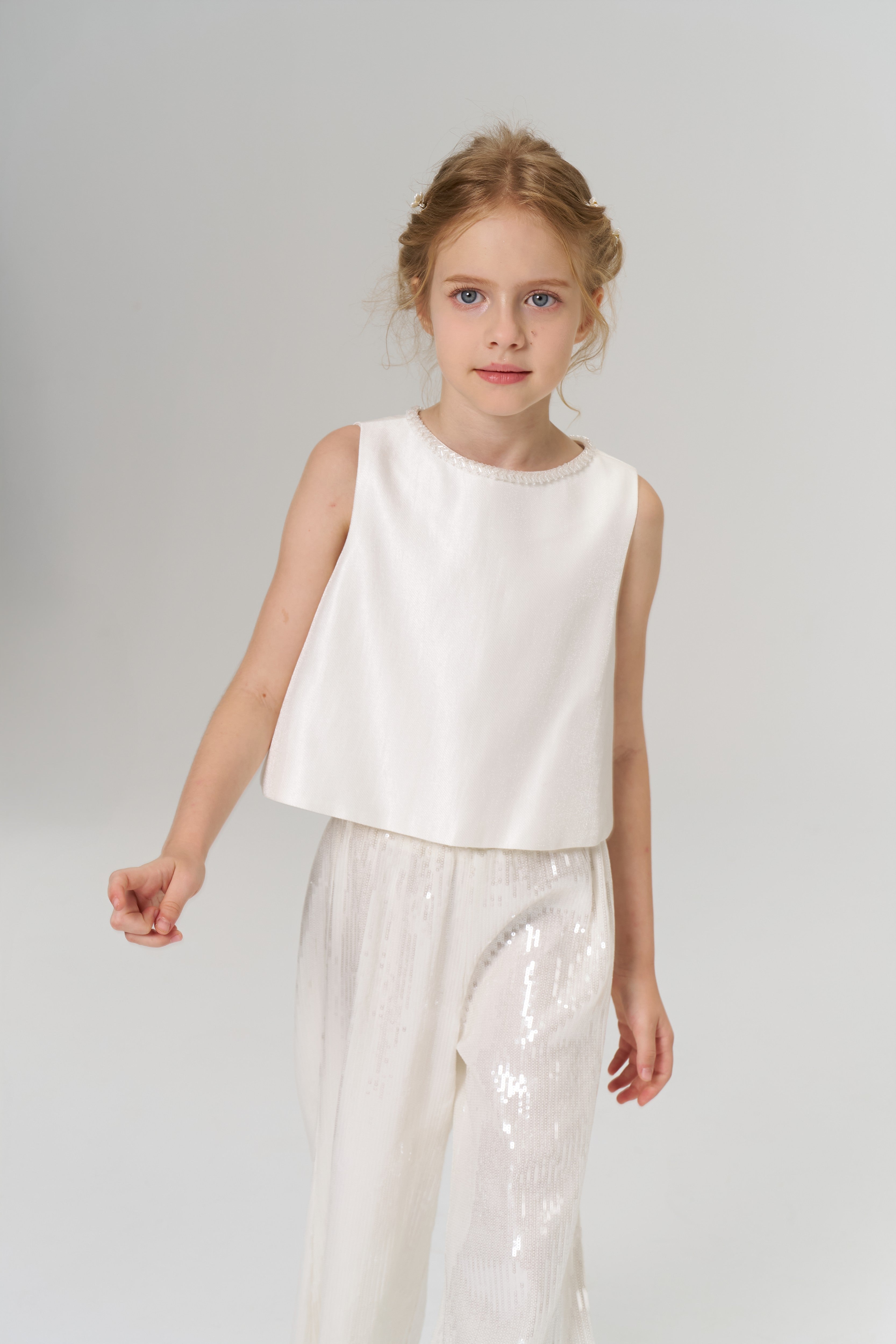 A young girl with light hair stands against a plain background, wearing the Radiance Satin Crop Top by KIKI and JOJO, featuring a hand-beaded pearl neckline, paired with shiny white pants.