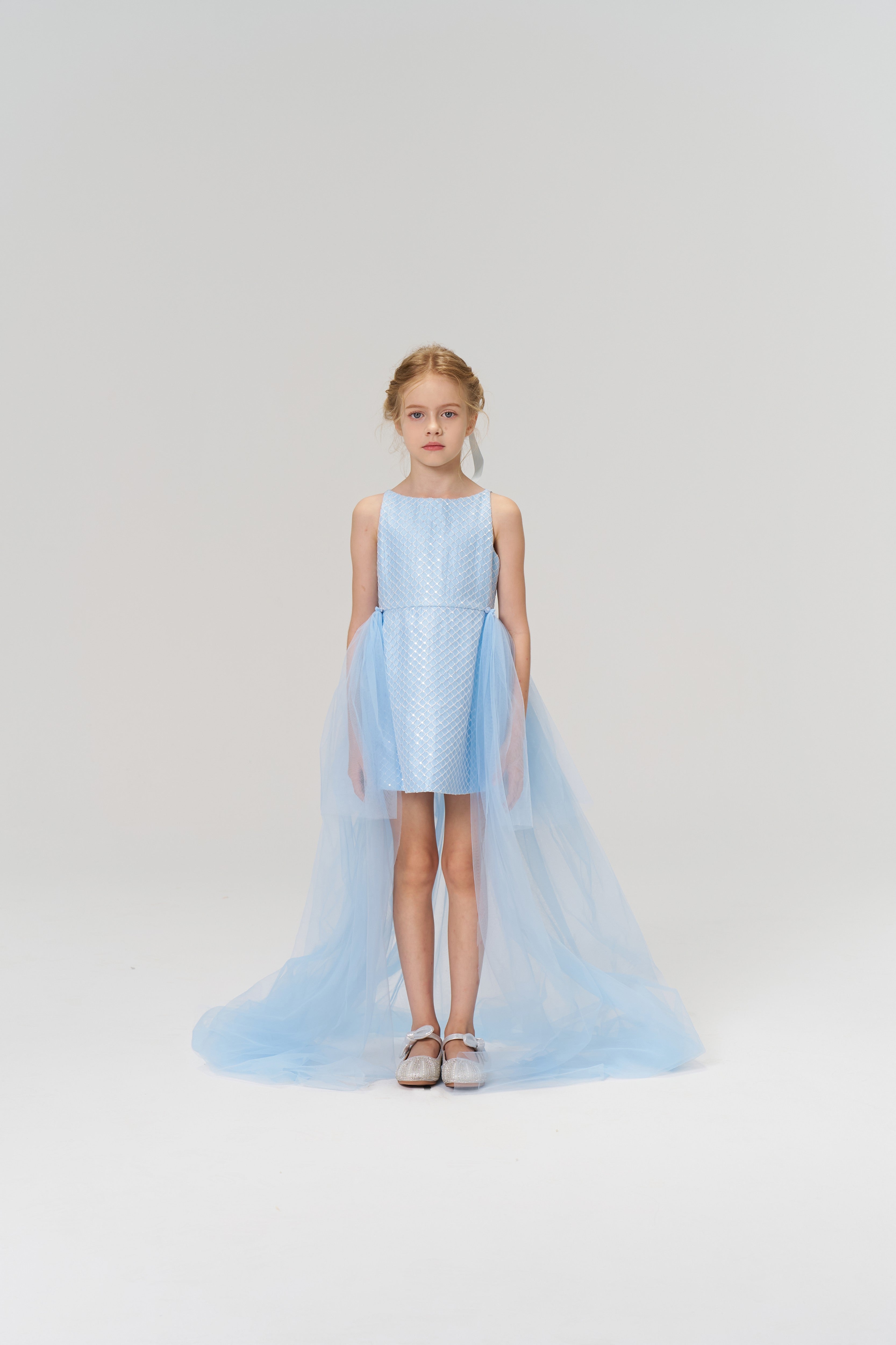 A young girl faces forward in the KIKI and JOJO Radiance Sequin Overlay Dress, featuring light blue tulle with sequins, a long train, and silver shoes, set against a plain white background.