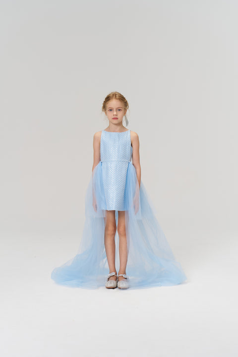 A young girl faces forward in the KIKI and JOJO Radiance Sequin Overlay Dress, featuring light blue tulle with sequins, a long train, and silver shoes, set against a plain white background.