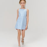 A young girl with blonde hair wears the KIKI and JOJO Radiance Sequin Overlay Dress in light blue and white shoes, standing against a plain white background.