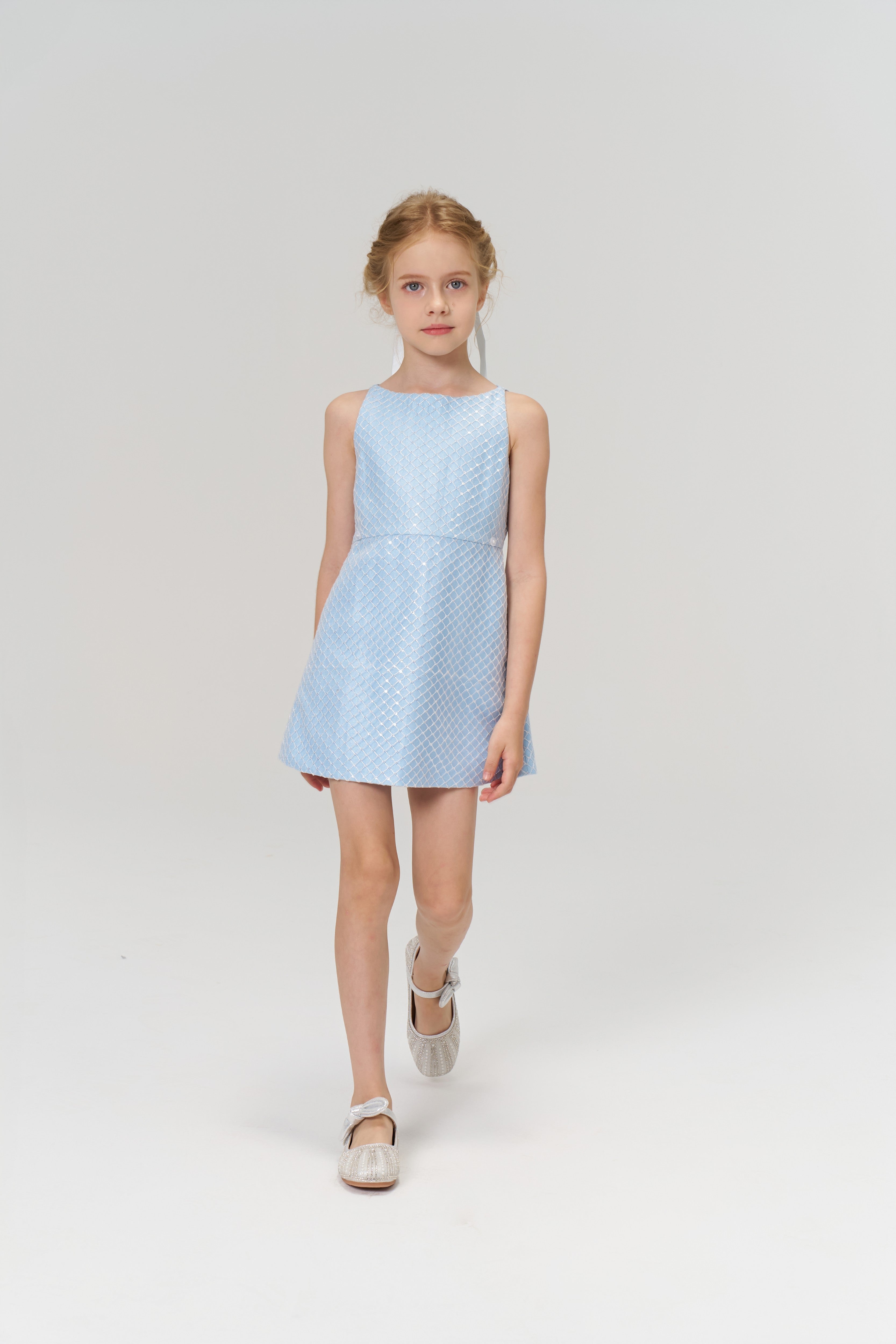 A young girl with blonde hair wears the KIKI and JOJO Radiance Sequin Overlay Dress in light blue and white shoes, standing against a plain white background.