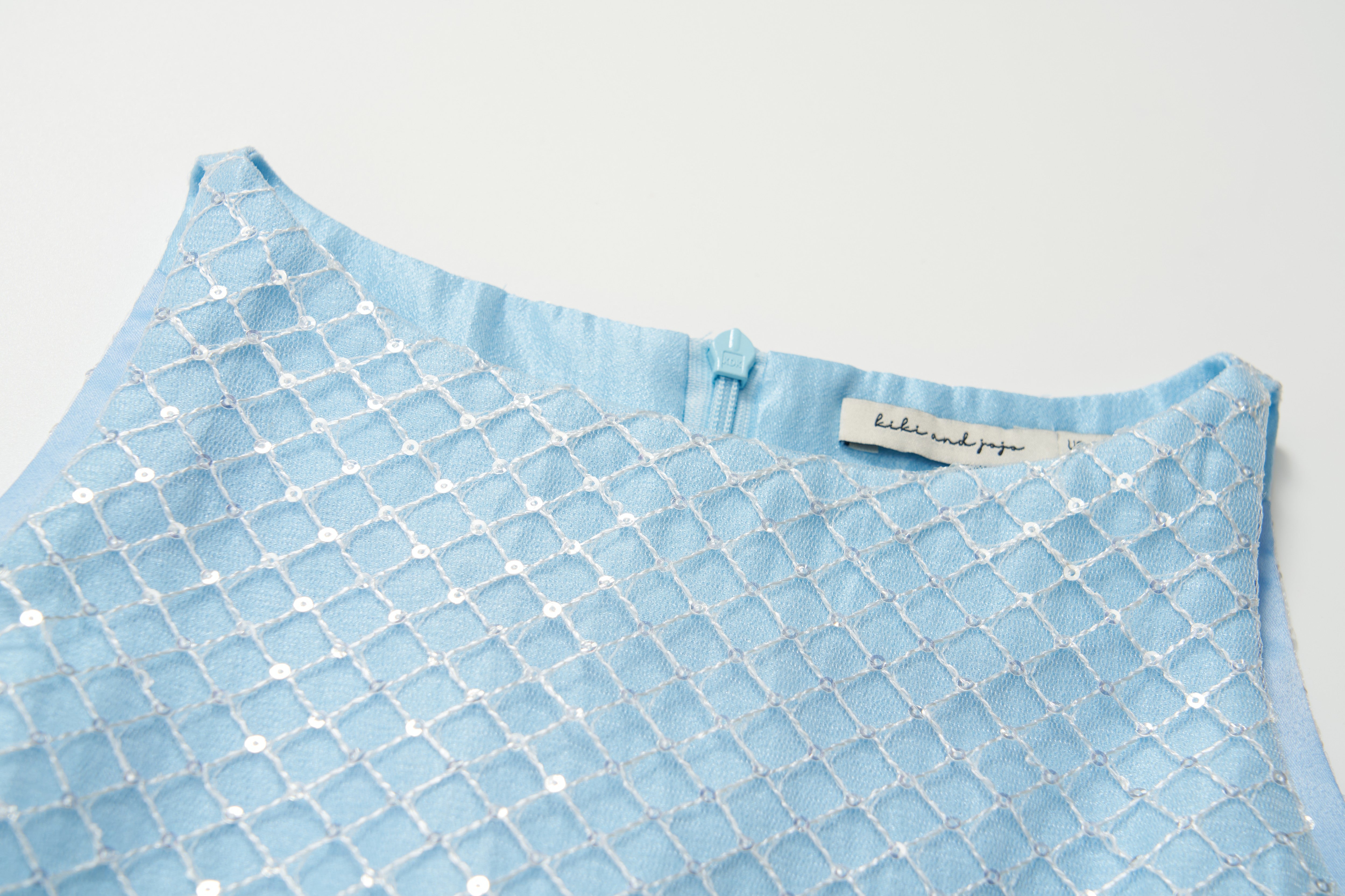 Close-up of the Radiance Sequin Overlay Dress by KIKI and JOJO, a light blue sleeveless formal dress featuring white embroidered geometric patterns, a back zipper, and a visible label inside the neckline.