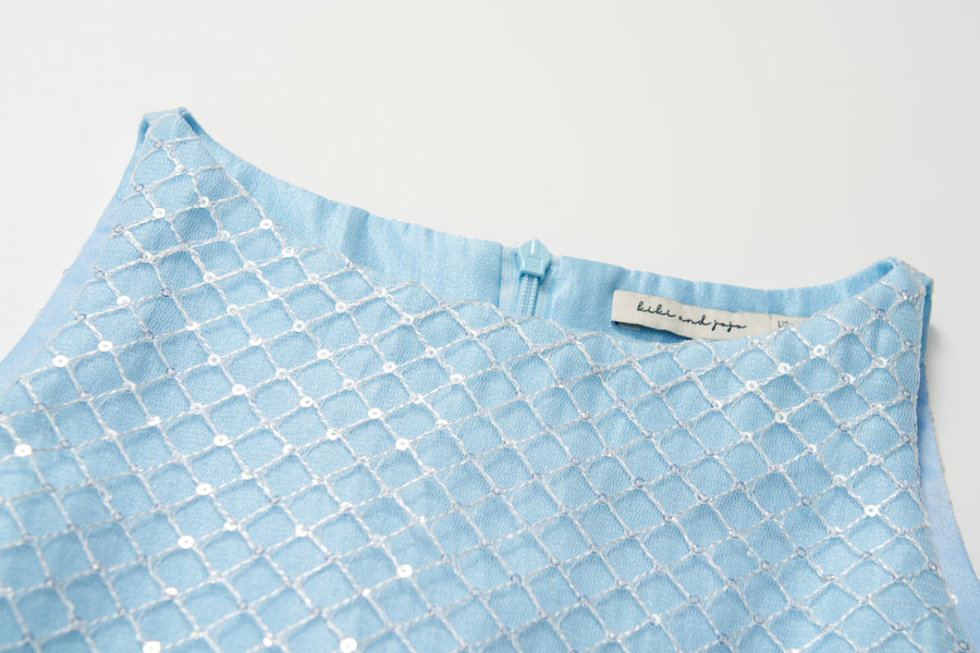 Close-up of the Radiance Sequin Overlay Dress by KIKI and JOJO, a light blue sleeveless formal dress featuring white embroidered geometric patterns, a back zipper, and a visible label inside the neckline.