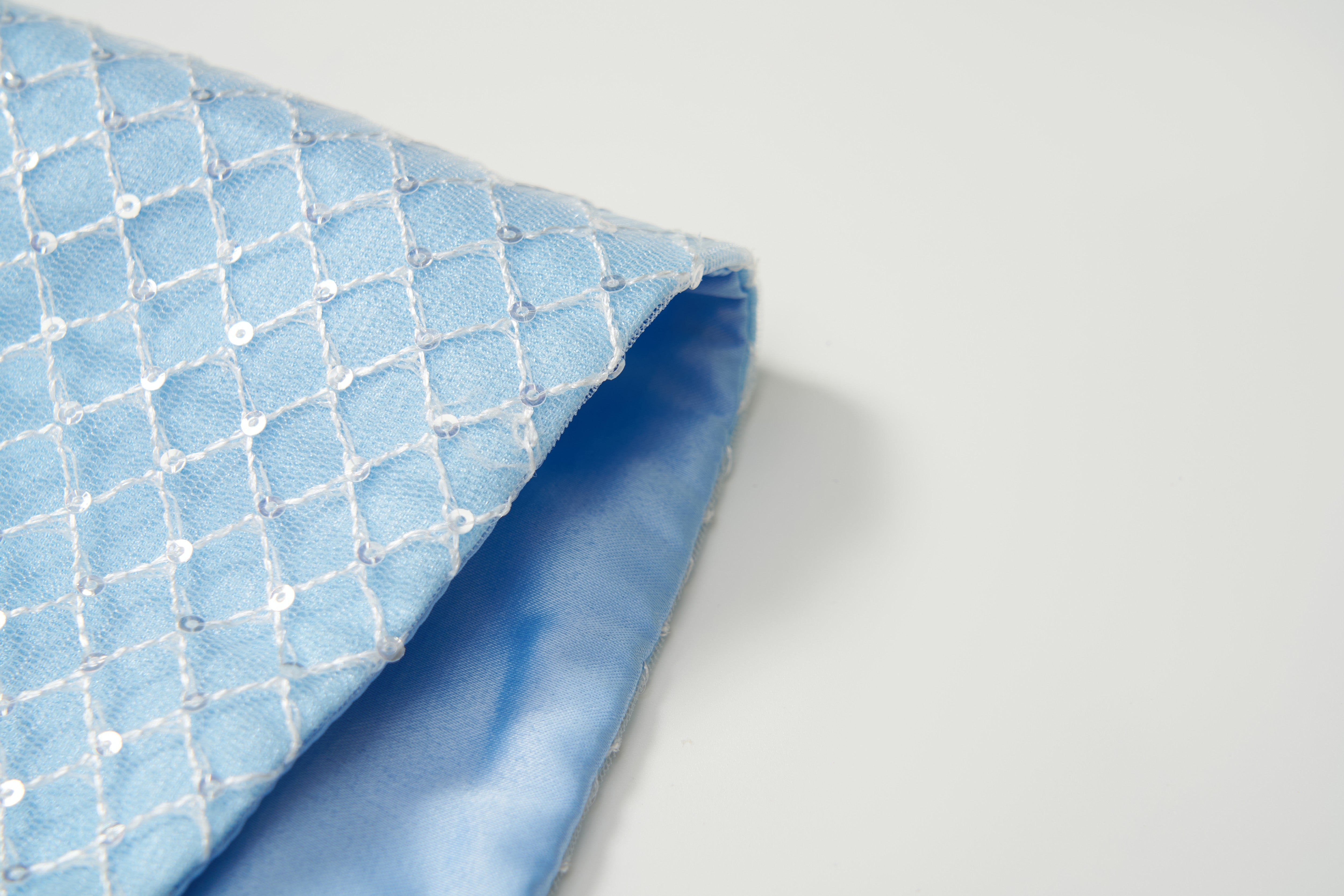 The KIKI and JOJO Radiance Sequin Overlay Dress features light blue diamond-patterned sequin tulle with a smooth, shiny blue lining—perfect for formal or convertible looks—displayed on a white surface.