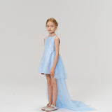 A young girl models the KIKI and JOJO Radiance Sequin Overlay Dress, featuring a light blue hue and a long sequin tulle train, as she poses against a plain white background.