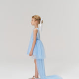 A young girl in profile wears the KIKI and JOJO Radiance Sequin Overlay Dress—a light blue tulle gown with a long train—and silver shoes, set against a plain white background.
