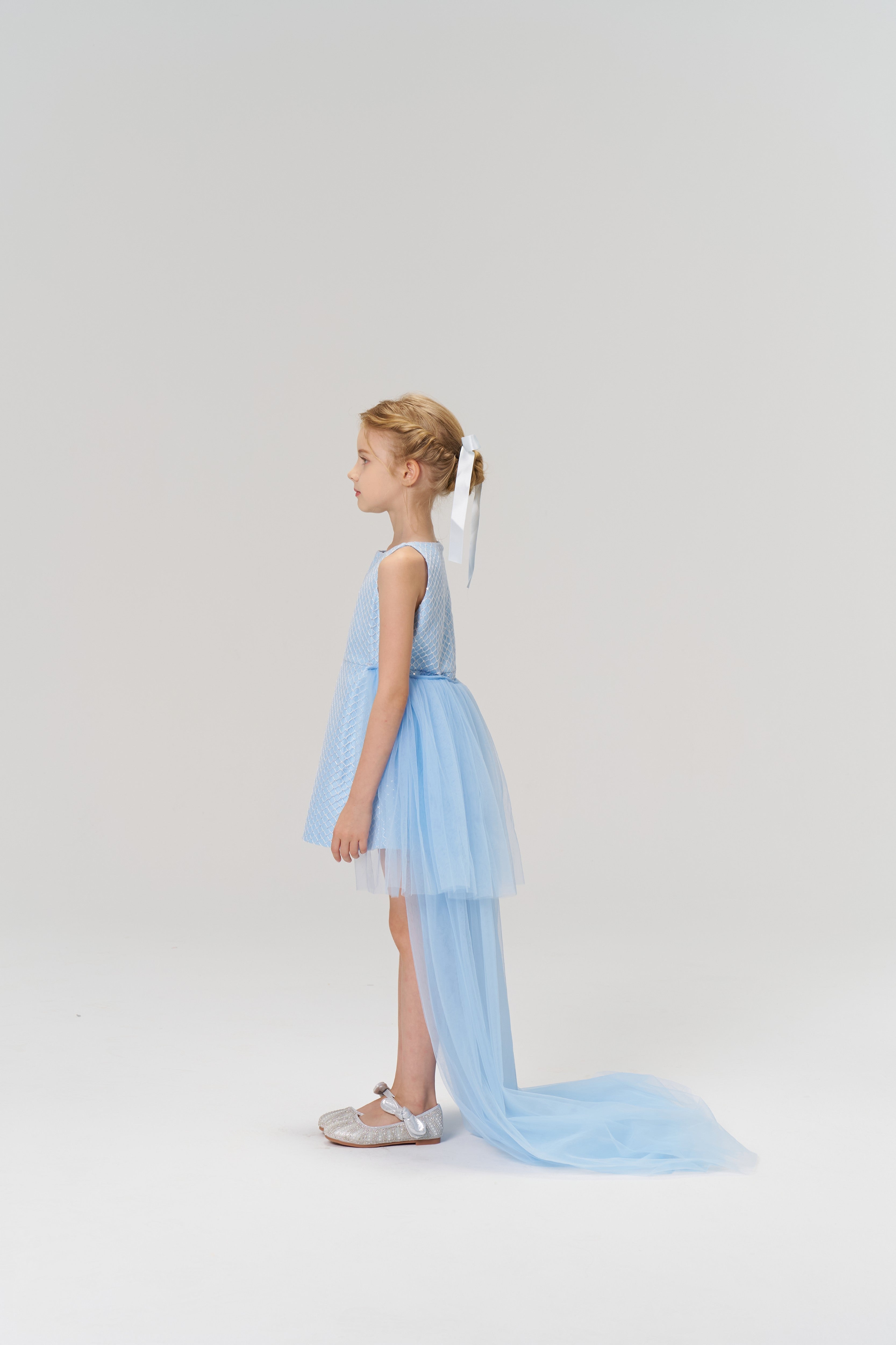 A young girl in profile wears the KIKI and JOJO Radiance Sequin Overlay Dress—a light blue tulle gown with a long train—and silver shoes, set against a plain white background.