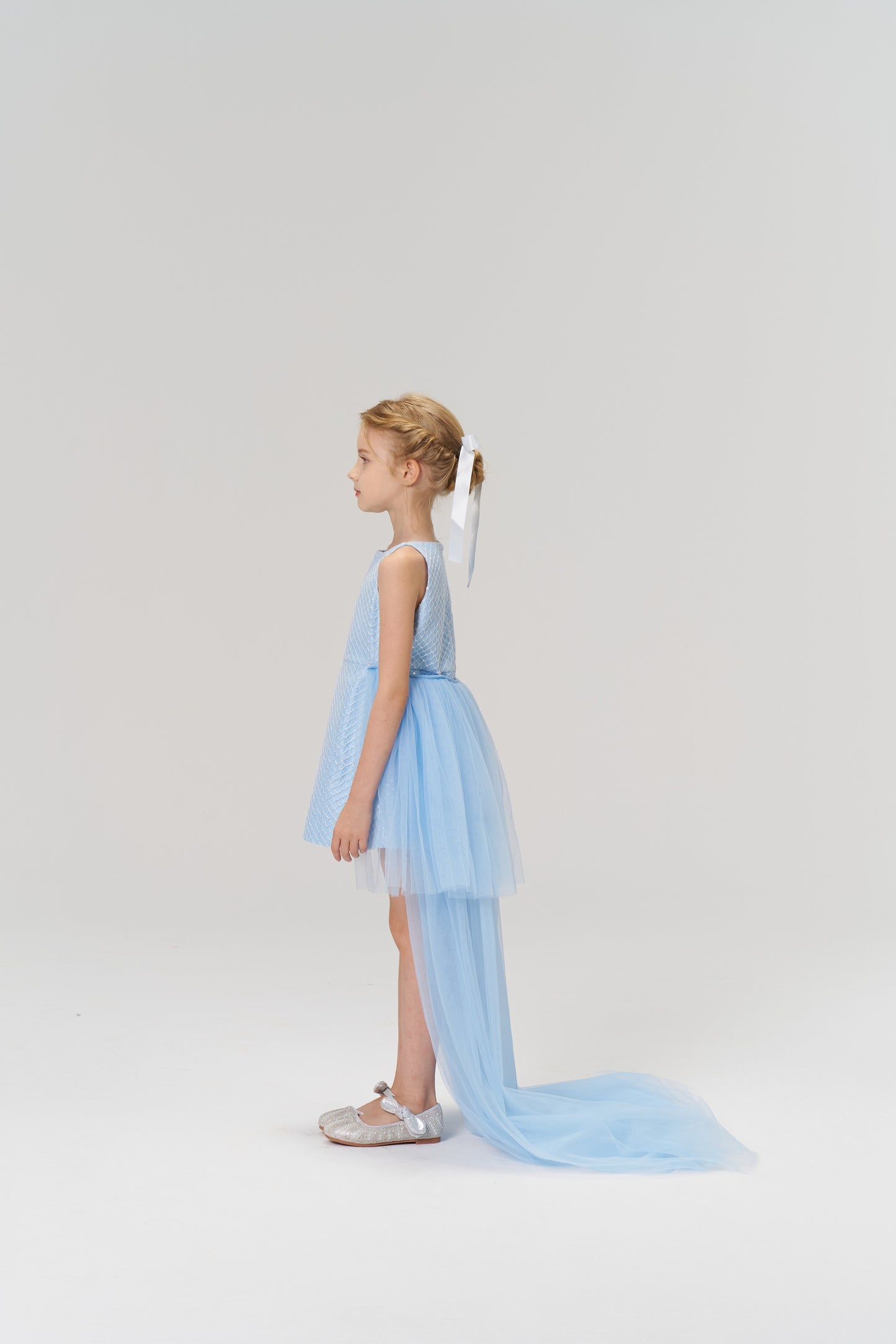 A young girl in profile wears the KIKI and JOJO Radiance Sequin Overlay Dress—a light blue tulle gown with a long train—and silver shoes, set against a plain white background.
