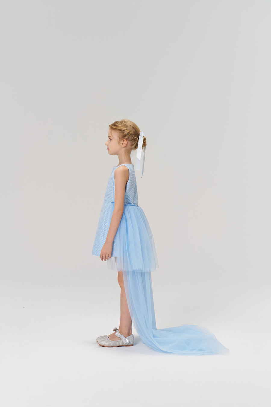 A young girl in profile wears the KIKI and JOJO Radiance Sequin Overlay Dress—a light blue tulle gown with a long train—and silver shoes, set against a plain white background.