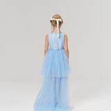 A young girl faces away in the KIKI and JOJO Radiance Sequin Overlay Dress, a light blue sleeveless tulle design, with a white ribbon in her hair, set against a plain light background.