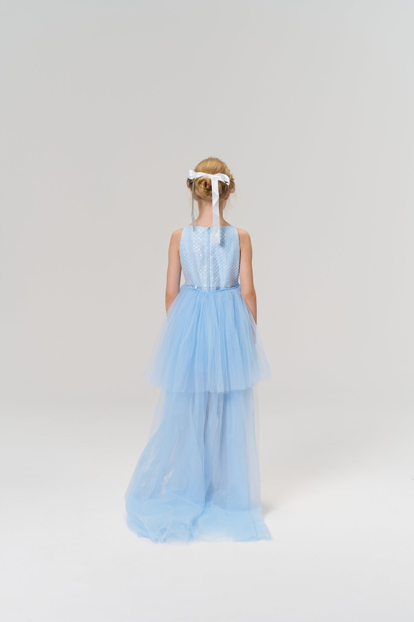 A young girl faces away in the KIKI and JOJO Radiance Sequin Overlay Dress, a light blue sleeveless tulle design, with a white ribbon in her hair, set against a plain light background.