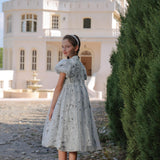A young girl wears the KIKI and JOJO Girls Cheongsam Dress – Ethereal Blossom Puff Sleeve Tulle, standing on a cobblestone path and looking back, with a large white house and tall greenery behind her.