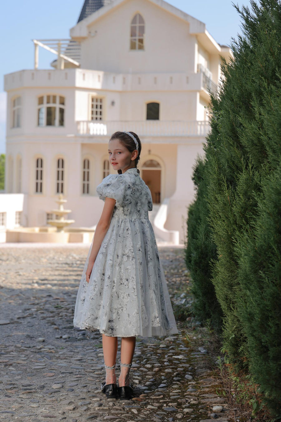 A young girl wears the KIKI and JOJO Girls Cheongsam Dress – Ethereal Blossom Puff Sleeve Tulle, standing on a cobblestone path and looking back, with a large white house and tall greenery behind her.