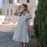A girl wears the KIKI and JOJO Girls Cheongsam Dress – Ethereal Blossom Puff Sleeve Tulle with black shoes, standing on a cobblestone path by a tall green hedge and a large white building in the background.