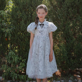 A young girl faces the camera outdoors in front of green shrubs, wearing the KIKI and JOJO Girls Cheongsam Dress – Ethereal Blossom Puff Sleeve Tulle.