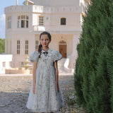 A young girl in the KIKI and JOJO Girls Cheongsam Dress – Ethereal Blossom Puff Sleeve Tulle stands on a cobblestone path before a white house with tall windows and a pointed roof.