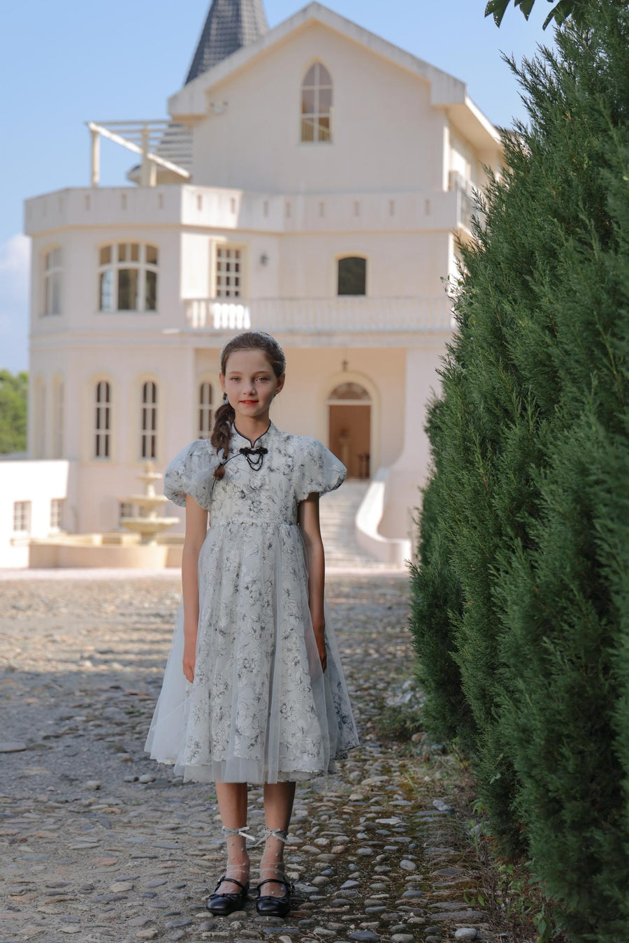 A young girl in the KIKI and JOJO Girls Cheongsam Dress – Ethereal Blossom Puff Sleeve Tulle stands on a cobblestone path before a white house with tall windows and a pointed roof.