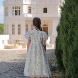 A girl wearing the KIKI and JOJO Girls Cheongsam Dress – Ethereal Blossom Puff Sleeve Tulle stands on a cobblestone path facing a large white house, with trees to the right.