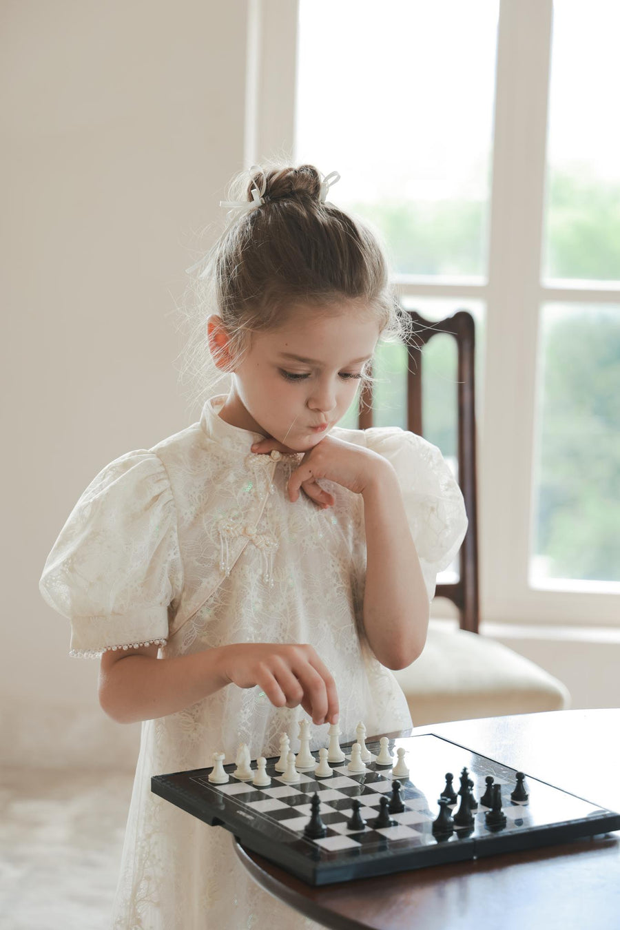A young girl in the KIKI and JOJO Puff Sleeve Tulle Cheongsam Dress with Pearl-Embroidered Cuffs stands indoors, thoughtfully playing chess at a small board.