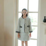A young woman stands indoors by a tall arched window, wearing the KIKI and JOJO Girls Tweed Jacket – Black & White Checkered Pearl with a matching skirt, paired with black shoes and socks.
