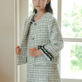 A young girl stands indoors by a window, wearing the KIKI and JOJO Girls Tweed Jacket – Black & White Checkered Pearl, paired with a matching skirt and headband.