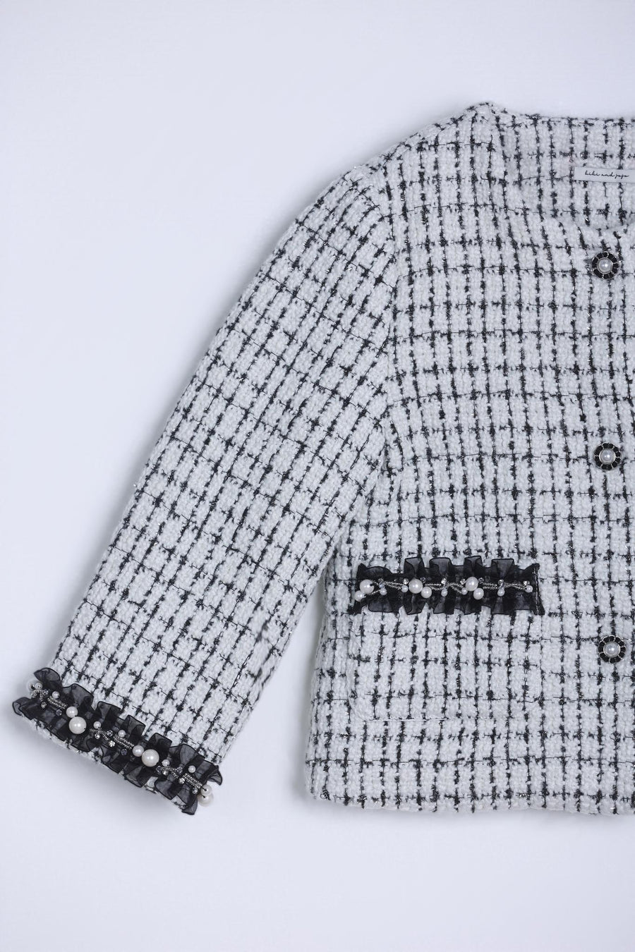A close-up of the KIKI and JOJO Girls Tweed Jacket in black-and-white checkered tweed, featuring decorative black trim and pearl accents on the sleeves and pockets.