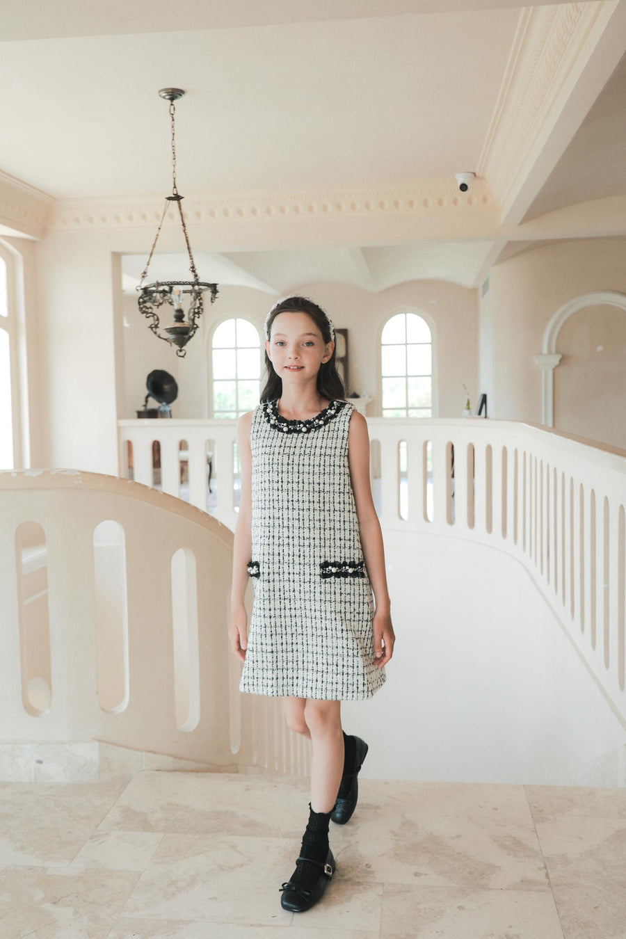 A girl in the KIKI and JOJO Girls Tweed Dress – Black & White Checkered Pearl stands on a marble staircase with arched windows behind her, capturing a timeless moment of luxury fashion.