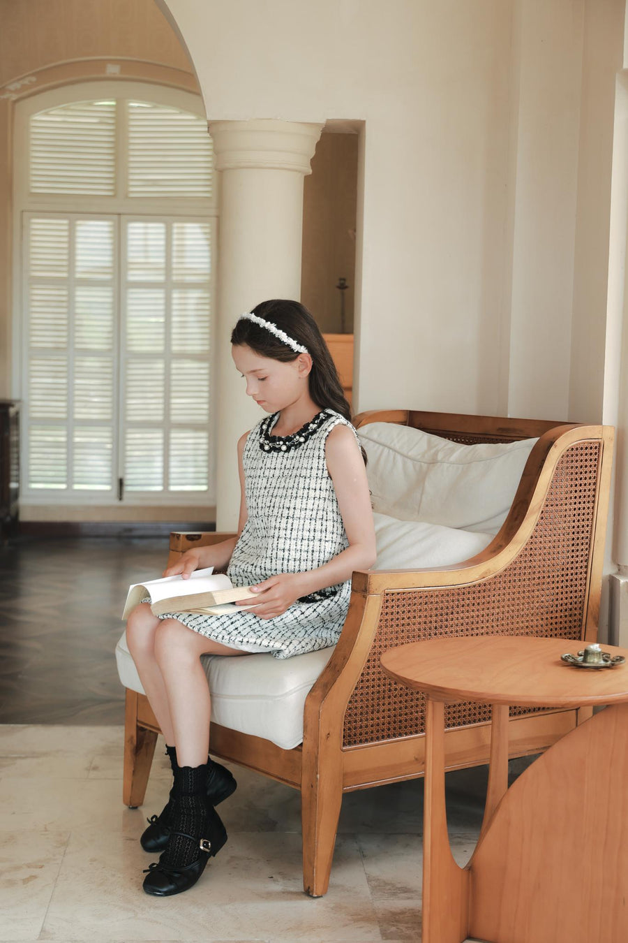 A young girl wearing the KIKI and JOJO Girls Tweed Dress – Black & White Checkered Pearl sits on a cushioned wooden chair indoors, reading a book, with a small wooden table beside her.