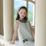 A young girl in the KIKI and JOJO Girls Tweed Dress – Black & White Checkered Pearl sits on a ledge between columns, with a headband, window, and greenery in the background.