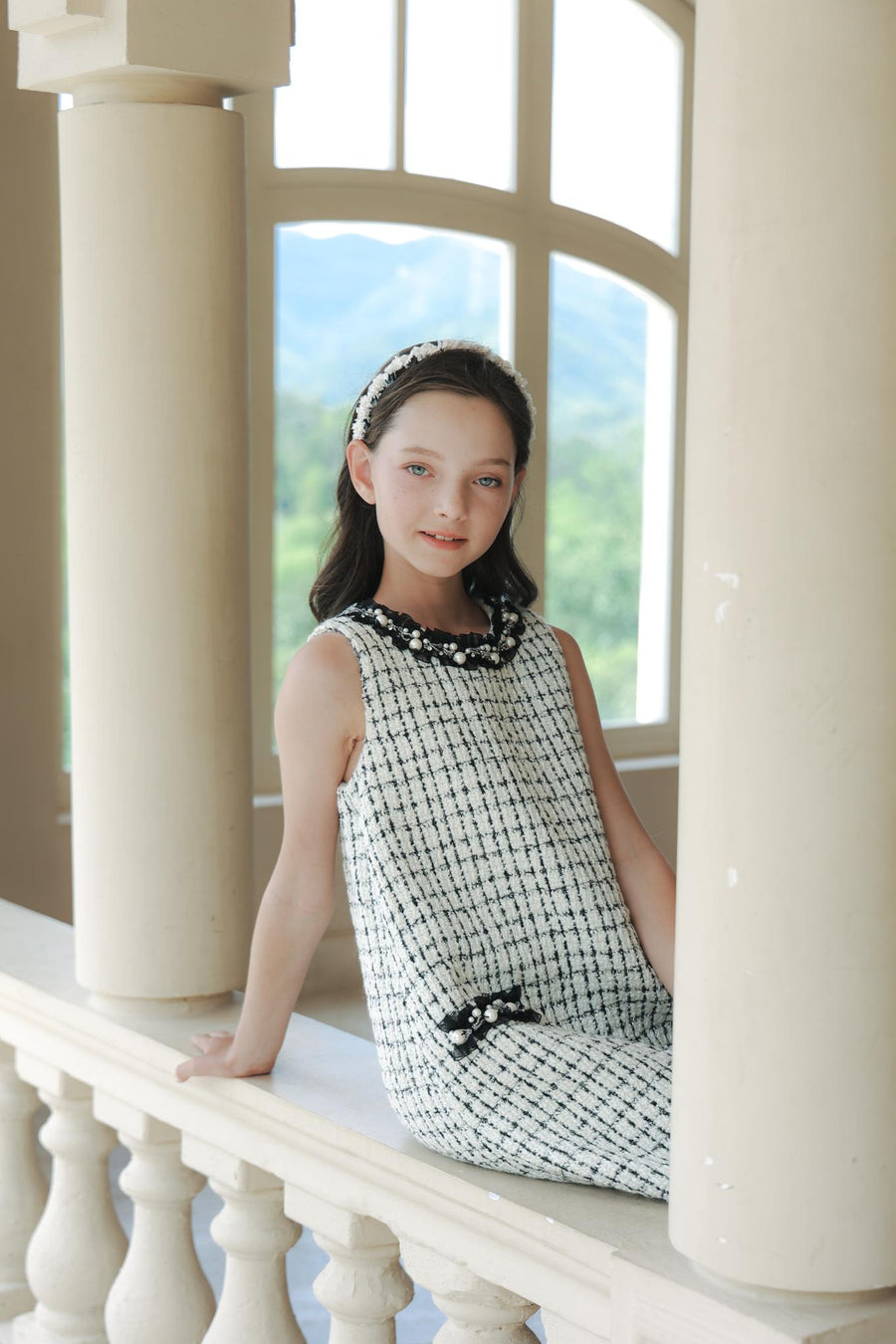 A young girl in the KIKI and JOJO Girls Tweed Dress – Black & White Checkered Pearl sits on a ledge between columns, with a headband, window, and greenery in the background.