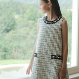A girl in a KIKI and JOJO Girls Tweed Dress – Black & White Checkered Pearl stands by a window, gazing at a green landscape with mountains in the background.