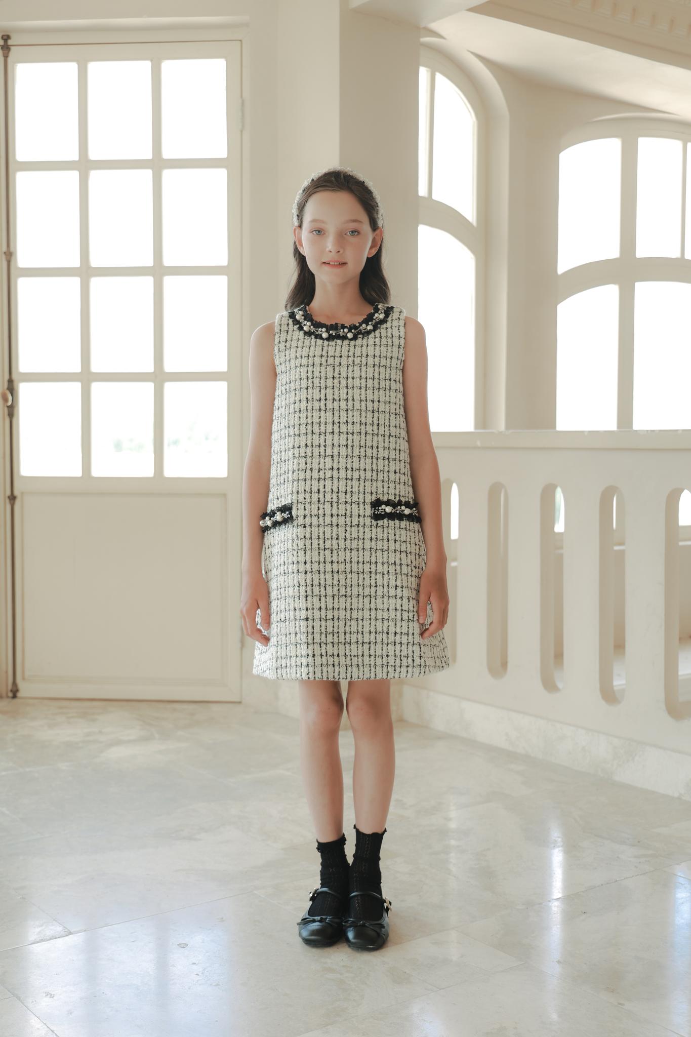 A girl stands indoors on a marble floor, wearing the KIKI and JOJO Girls Tweed Dress – Black & White Checkered Pearl, paired with black socks and shoes, exuding timeless luxury fashion.