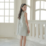 A girl with long dark hair stands indoors, looking back over her shoulder, wearing the KIKI and JOJO Girls Tweed Dress in Black & White Checkered Pearl with black shoes.