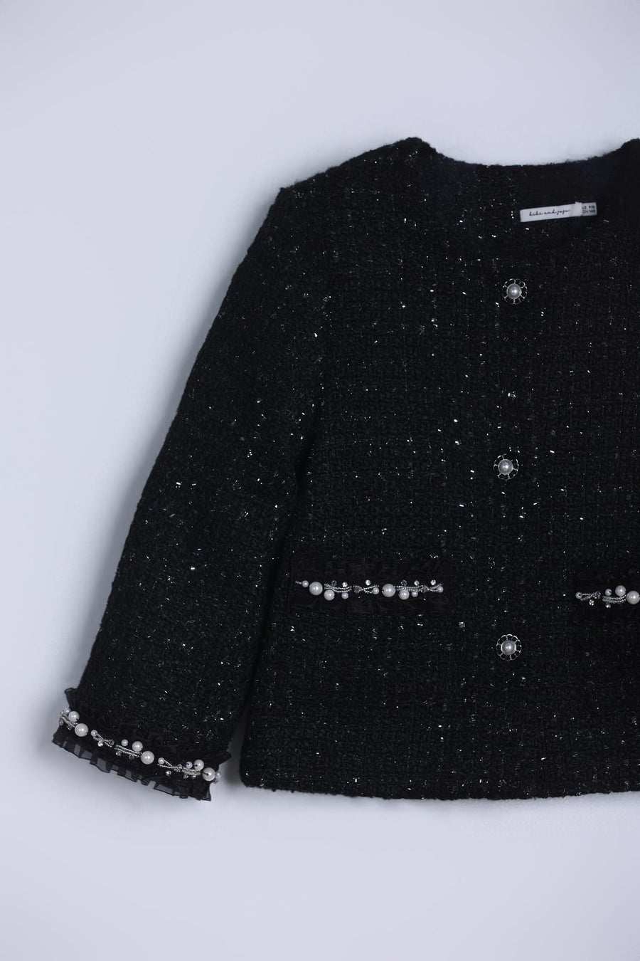 The Girls Tweed Jacket – Classic Black Pearl by KIKI and JOJO features long sleeves, front buttons, and vintage-inspired hand-beaded pearl details on the cuffs and pockets, shown against a light background.