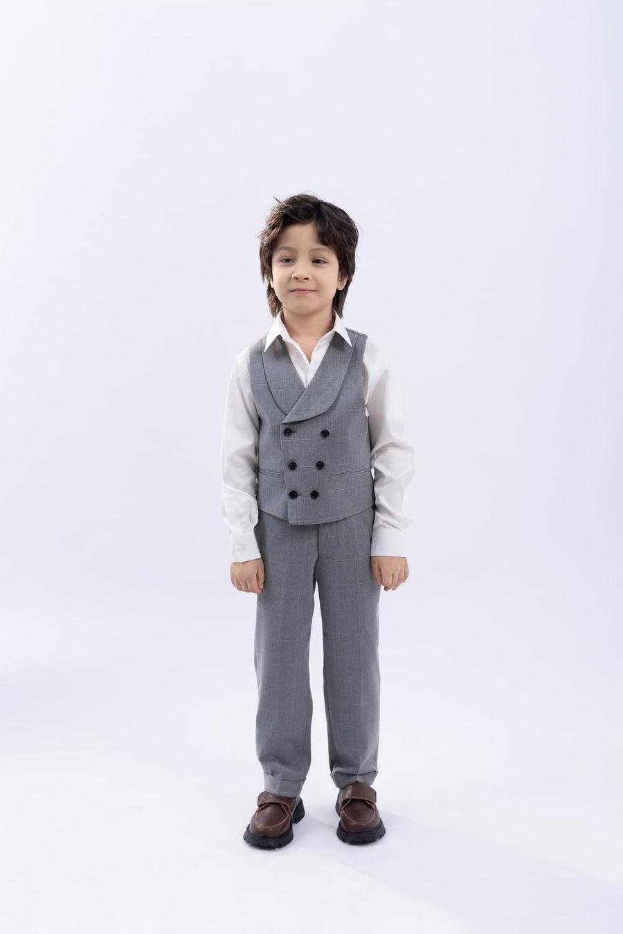 A young boy faces forward in a KIKI and JOJO Kids Suit Jacket – Light Gray, checkered and wrinkle free, paired with a double-breasted vest, white shirt, and brown shoes against a plain light background.