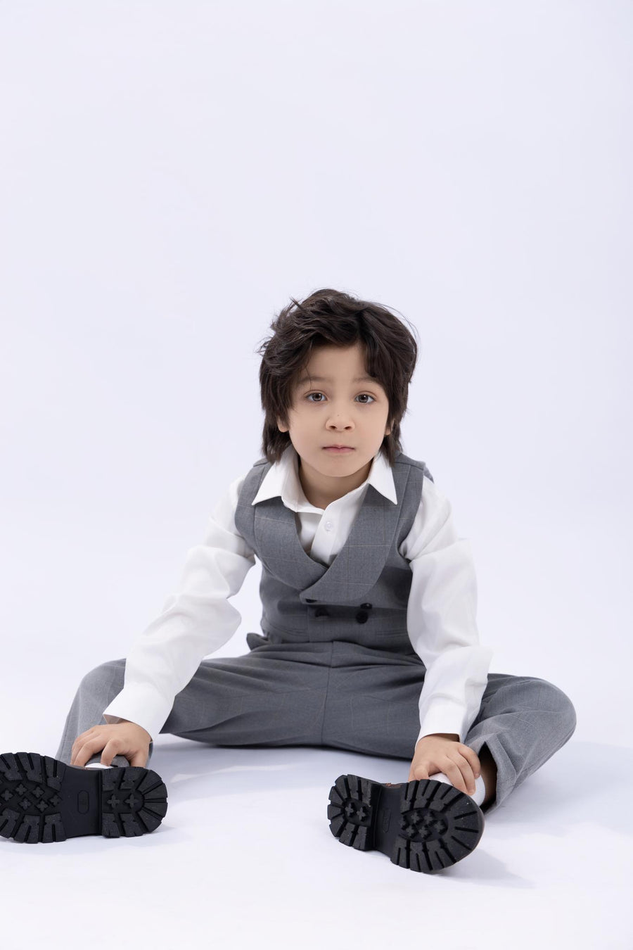 A young boy wears the KIKI and JOJO Kids Suit Jacket – Light Gray (3-14 Years), checkered and wrinkle free, over a white shirt, sitting on the floor with legs apart against a plain white background.