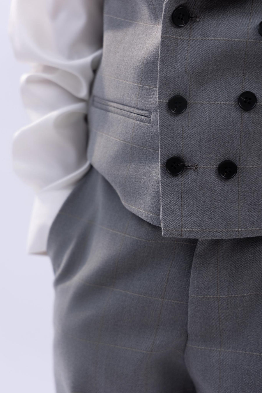 A child wears the KIKI and JOJO Kids Suit Jacket in light gray checkered, wrinkle-free fabric, shown waist to mid-chest with black buttons, white shirt, and one hand in pocket—stays sharp all day.