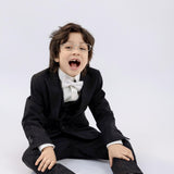 A young boy in glasses, black shoes, and white socks sits on the floor wearing KIKI and JOJO Kids Suit Pants – Black (3-14 Years), classic fit and wrinkle-free, with a modern flat-front design against a plain light background.