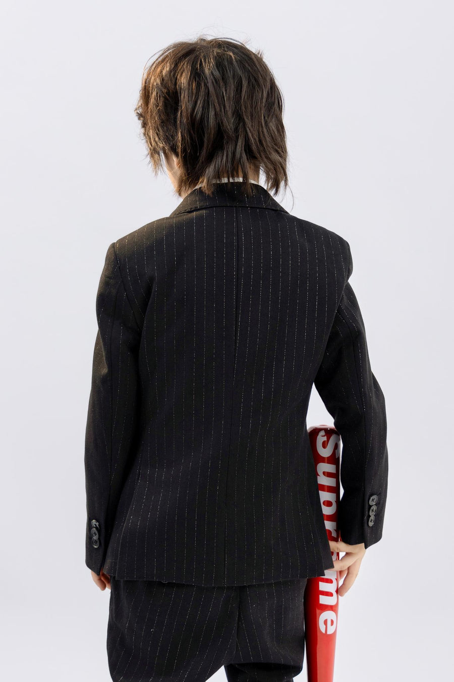 A child in KIKI and JOJO Kids Suit Pants – Black (3-14 Years), classic fit and wrinkle-free, stands with their back to the camera, holding a red object labeled "Supreme" behind them.