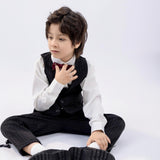 A young boy in a suit and bow tie sits on the floor, one hand on his chest, wearing KIKI and JOJO Kids Suit Pants – Black (3-14 Years), Classic Fit, Wrinkle-Free, looking to the side against a plain white background.