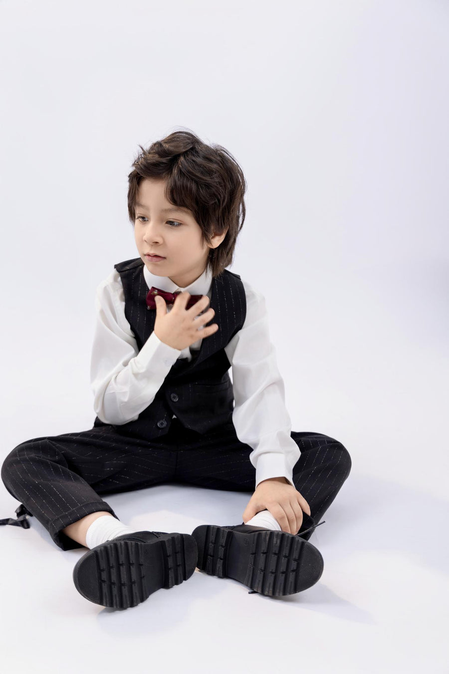 A young boy in a suit and bow tie sits on the floor, one hand on his chest, wearing KIKI and JOJO Kids Suit Pants – Black (3-14 Years), Classic Fit, Wrinkle-Free, looking to the side against a plain white background.