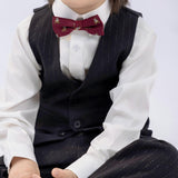 A young child in a white shirt, black pinstripe vest, red bow tie, and KIKI and JOJO Kids Suit Pants – Black (Classic Fit, Wrinkle-Free) is sitting cross-legged with their mouth open, possibly speaking or singing.