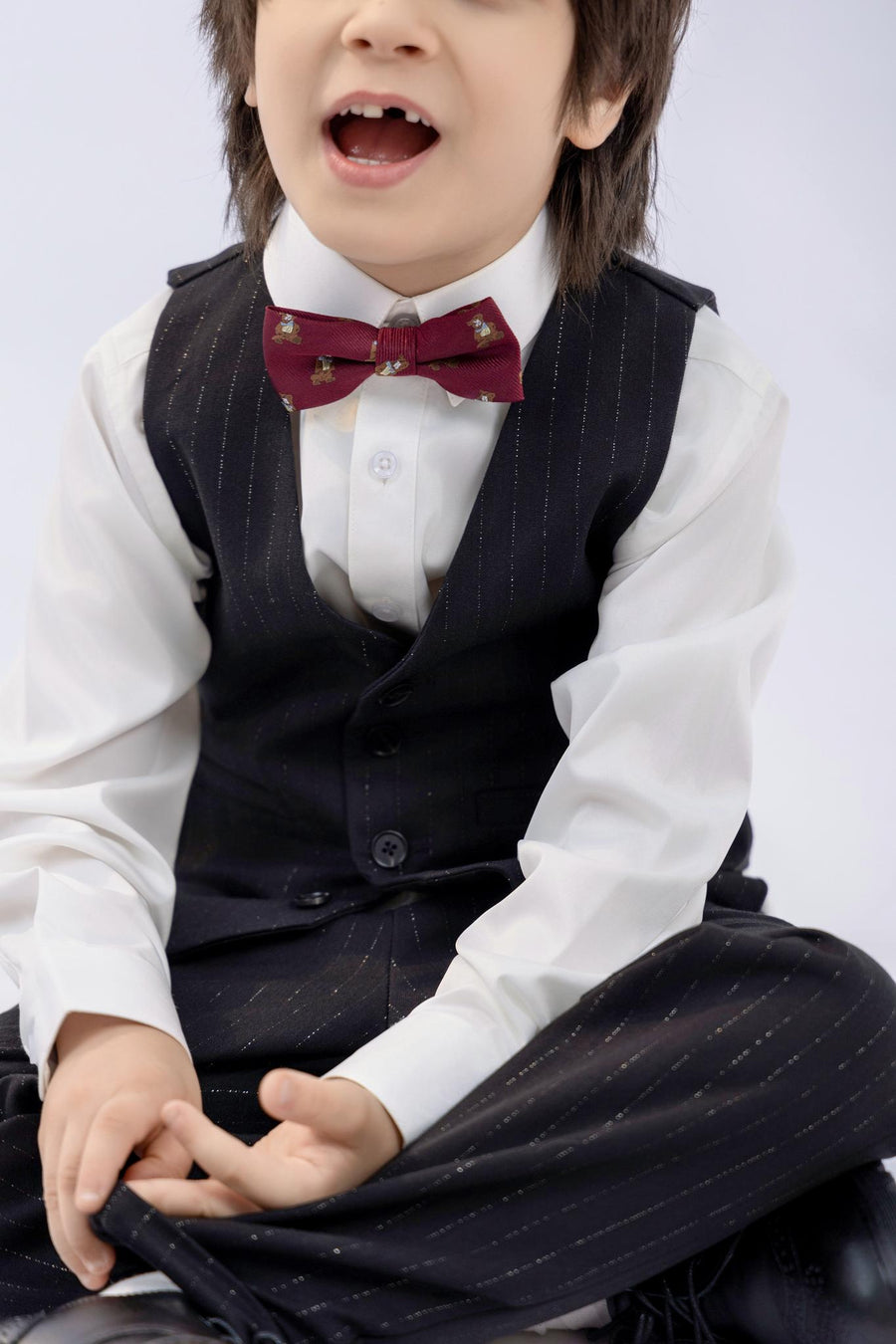 A young child in a white shirt, black pinstripe vest, red bow tie, and KIKI and JOJO Kids Suit Pants – Black (Classic Fit, Wrinkle-Free) is sitting cross-legged with their mouth open, possibly speaking or singing.