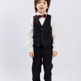 A young boy faces forward in KIKI and JOJO Kids Suit Pants – Black (3-14 Years), classic fit and wrinkle-free, paired with a white shirt, red bow tie, and black dress shoes, set against a plain light background.