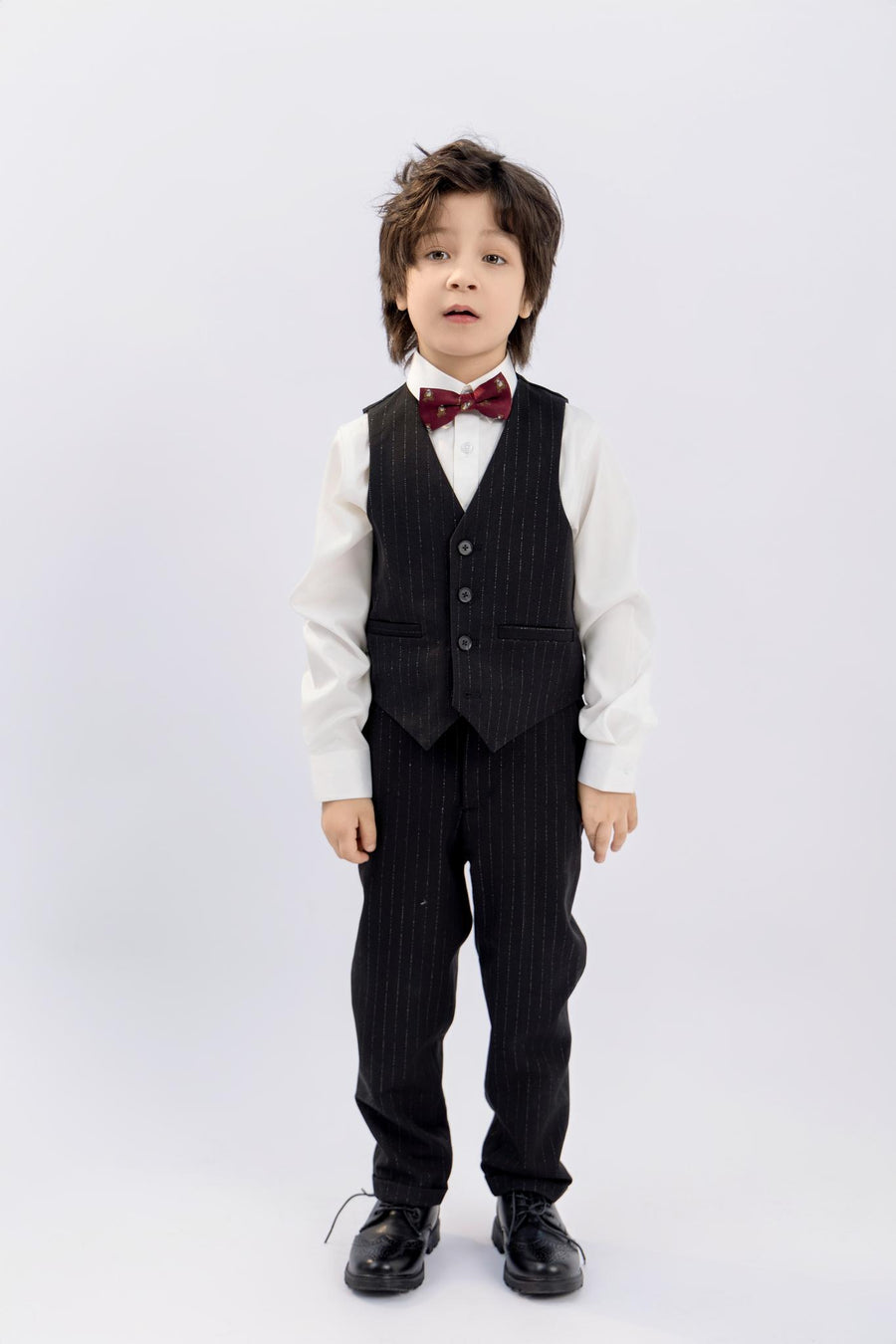 A young boy faces forward in KIKI and JOJO Kids Suit Pants – Black (3-14 Years), classic fit and wrinkle-free, paired with a white shirt, red bow tie, and black dress shoes, set against a plain light background.