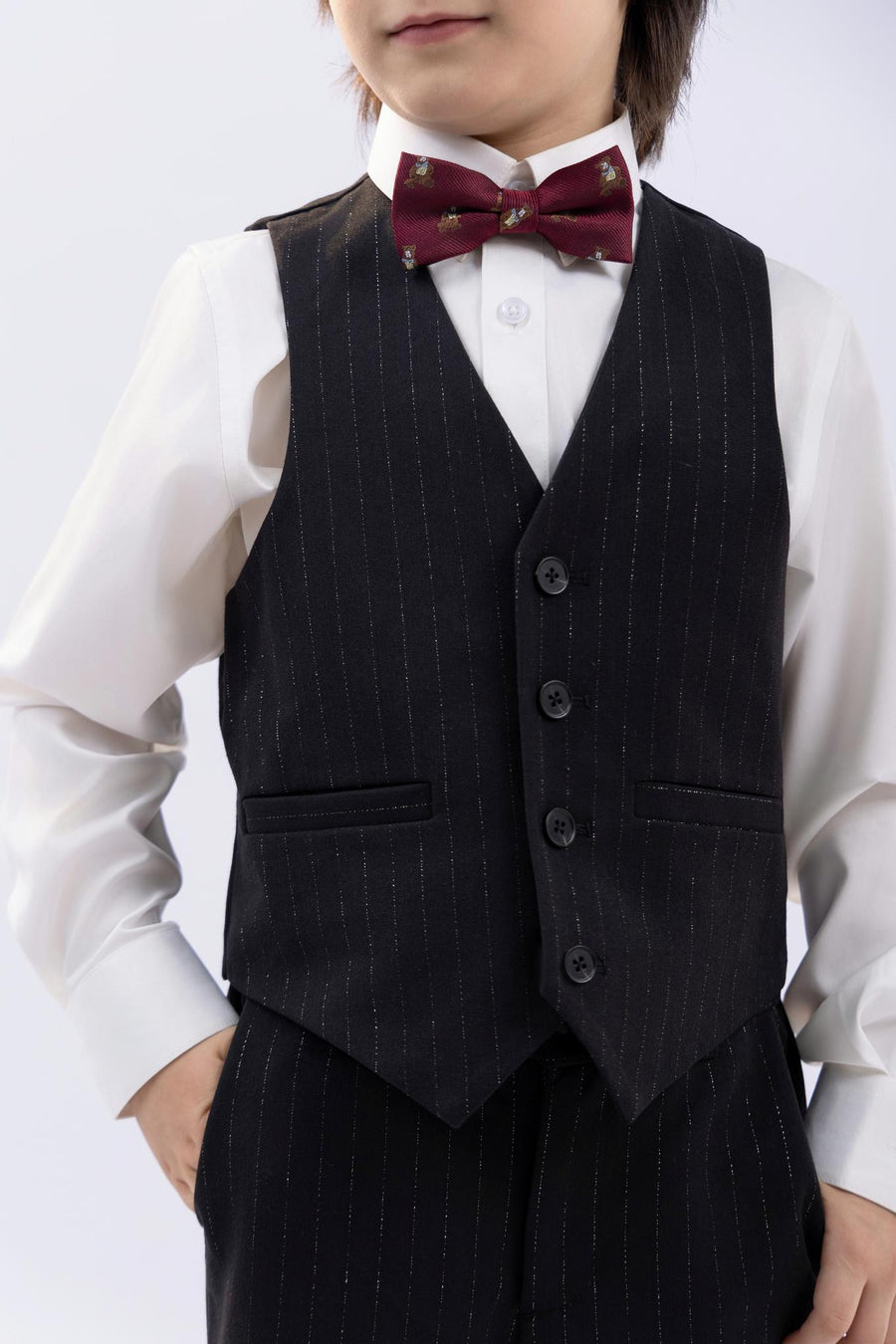 A child wears a white shirt, dark vest, maroon bow tie, and KIKI and JOJO Kids Suit Pants – Black (Classic Fit, Wrinkle-Free), with one hand in their pocket.
