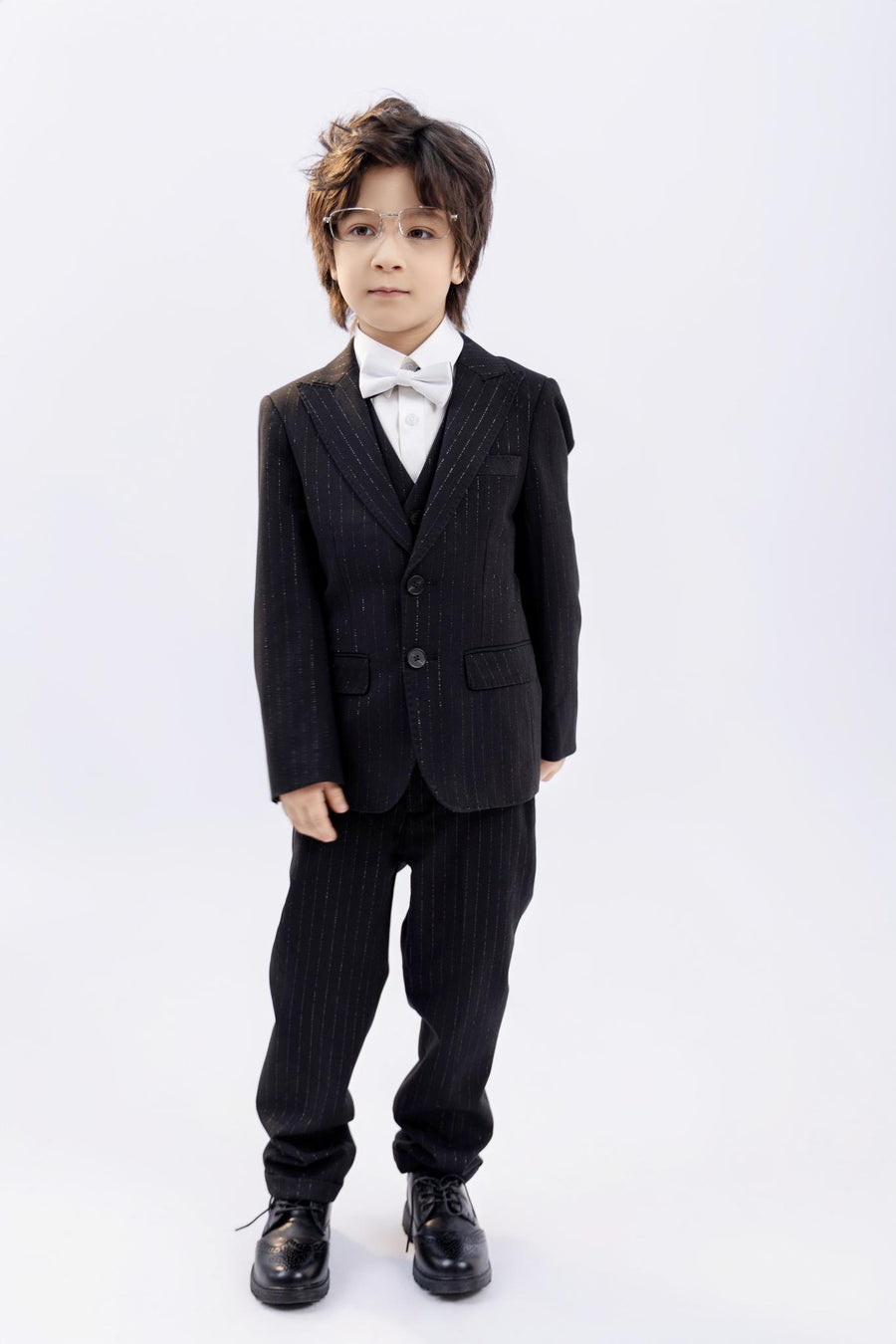 A young boy wears KIKI and JOJO Kids Suit Pants – Black (Classic Fit, Wrinkle-Free, 3-14 Years), paired with a white shirt and bow tie, standing against a plain white background.